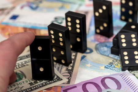 Finger pushing domino standing on euro and dollar cash.