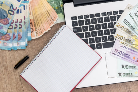 laptop, pen, euro banknote and notepad on desk.の写真素材