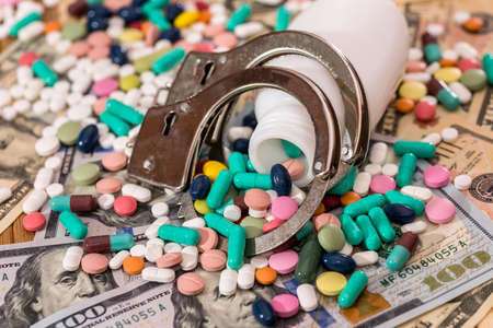 Dispersed pills from container with handcuffs on dollar banknotes