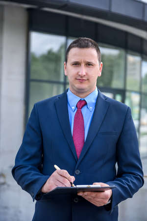 Young businessman in suit with clipboard standing outdoors