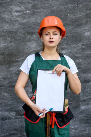 Woman in protective uniform and helmet posing with clipboard and tool beltの写真素材