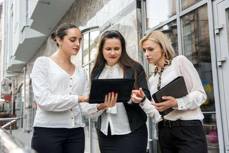 Business meeting outside building. Three young ladies in business suits discussing information on...