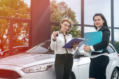 Two pretty women with folders standing near new car. They exam some documents in foldersの写真素材