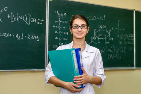 Chemist with folders standing and posing for camera. Behind woman university board with chemical...