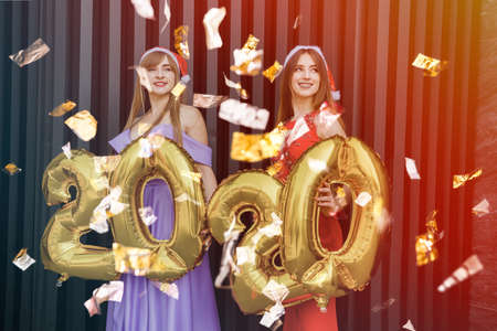 Portrait of two smiling attractive girls in elegant dresses posing while standing with gold colored 2020 number balloons and looking at cameraの写真素材