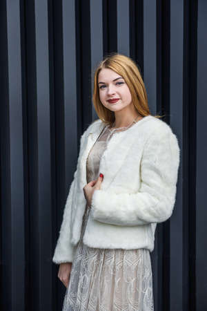 Fashion. Elegant woman in beige dress wearing in white fur coat. Beautiful girl model in luxurious clothes posing on abstract background. Outdoor closeup portrait.の写真素材