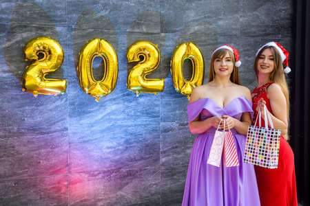 Two sisters in elegant dresses with presents posing with golden air balloons 2020 on abstract...