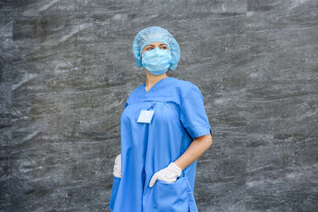 Medical nurse in potective uniform posing for camera. She's wearing blue mask, hat and shirtの写真素材