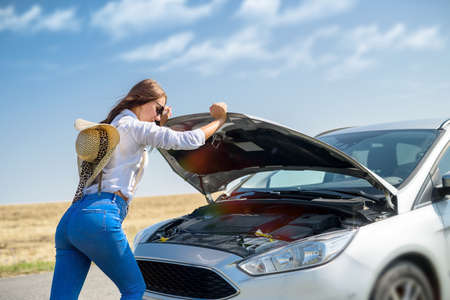 Pretty young woman looking at the engine of broken car. Road trip problems.