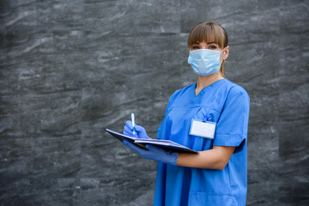 Doctor with notepad. Woman in blue medical coat posing on gray background in mask with stethoscopeの写真素材