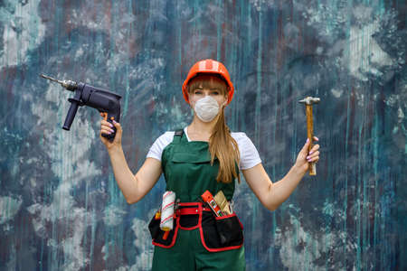 Girl in construction clothes and protective equipment posing with a drill and hammer on gray wall.の写真素材