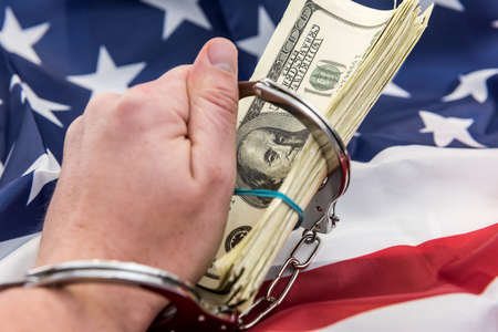Male hand in closed metal handcuff holding american dollars cash over flag of USA. Illegal concept. Bribing and corruptionの写真素材