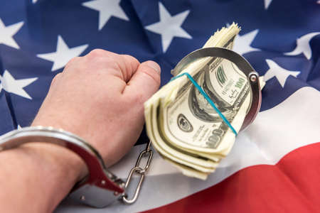 Male hand in closed metal handcuff holding american dollars cash over flag of USA. Illegal concept. Bribing and corruptionの写真素材