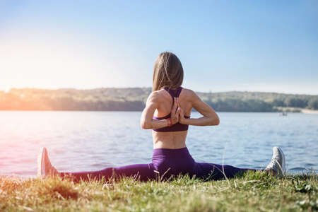 young and attractive woman in sportswear stretching warm-up twine on grass near the lake at daytime....