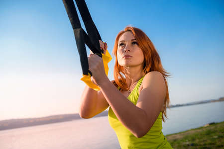 Young woman exercising with suspension trainer sling in park, near the lake.の写真素材