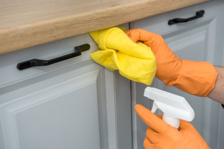 Male's hands in yellow rag glove with spray and sponge washing kitchen wooden furniture. cleaning...