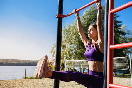 Portrait of strong young woman in sports clothing hanging on wall bars with her legs up. Fitness...