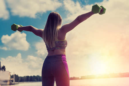 Young fitness girl making exercises with dumbbells on sunset near lake in city park