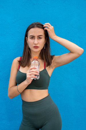 Pretty slim young woman in sports clothing with holding water bottle isolated on blue background. healthy lifestyleの写真素材