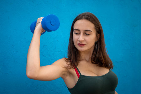 young woman in green sport cloth holding blue dumbbell isolated on blue. Health and beauty lifestyle