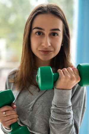 young beautiful woman doing exercise with dumbbells while standing in front of window at gymの写真素材