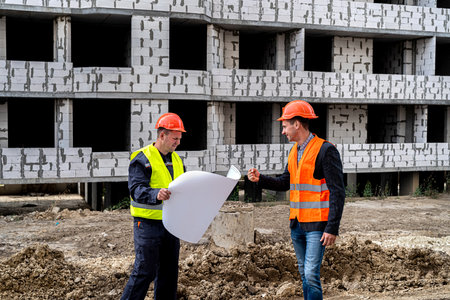 two young workers in uniform on a construction site stand with a plan and compare with the new...