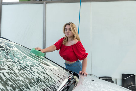 beautiful woman driver cleaning her car with green sponge in foam at manual auto washing stationの写真素材