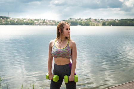 Young fitness female instructor doing workout with dumbbells by the river. healthy lifestyleの写真素材