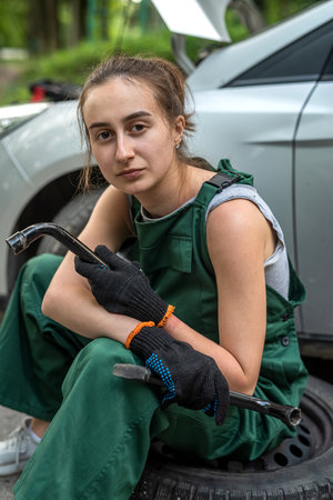 Young female mechanic in green uniform standing near a broken-down car and waiting help, outdoor