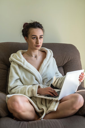 young successful girl in home clothes sitting on sofa with laptop working online during pandemic. The concept of work after a pandemicの写真素材