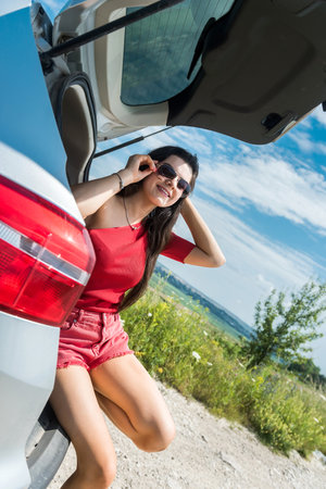 happy traveling woman sitting in car trunk and resting on stop in rural field. summer lifestyleの写真素材