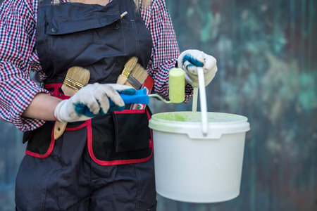 pretty woman in uniform and helmet holding paint and roller getting ready to renovate house. construction worker.の写真素材