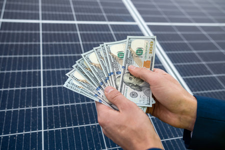 beautiful young hand in a blue jacket holds dollars near the new solar panels. green electricity conceptの写真素材