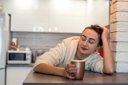 beautiful young girl with beautiful hair in a white coat drinking coffee in the kitchen in the morning. Sleepy morning conceptの写真素材