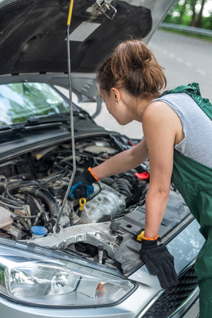 female mechanic wear green uniform checking level of oil on a car engine dipstick, outdoors.