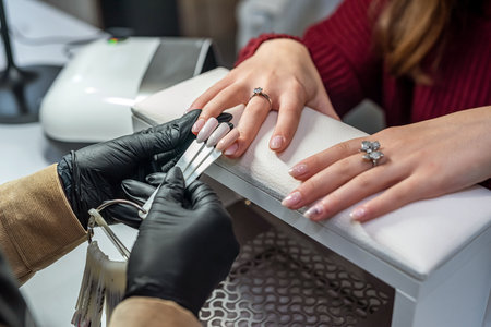 A beautiful young woman working in a nail salon offers a color of nail polish to one of her clients and smiles. The concept of the salon where you do manicuresの写真素材
