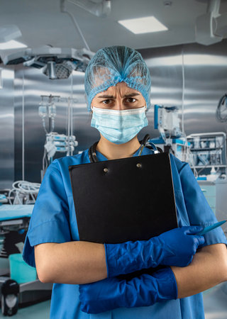 Female doctor in blue uniform and protective mask, gloves writes notes on the clipboard in operating room at hospitalの写真素材