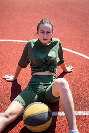 Attractive young slim woman posing sitting on a basketball court holding a basketball. urban lifestyle. hot summer day. sport for healthの写真素材