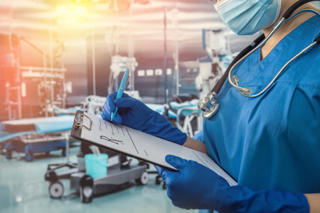 Female doctor in blue uniform and protective mask, gloves writes notes on the clipboard in operating room at hospitalの写真素材