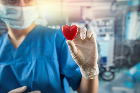 Female doctor or nurse in blue uniform, gloves mask holding small red heart in operating room at...