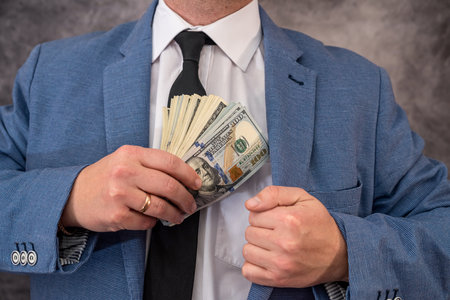 A man businessman member or officer in a suit puts a bribe in the form of hundred-dollar bills in his pocket. The concept of corruption and bribery among officialsの写真素材