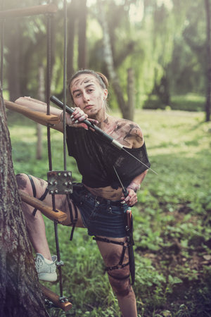 beautiful young girl in short shorts and a T-shirt smeared with swamp holds a dagger and a knife in the woodsの写真素材