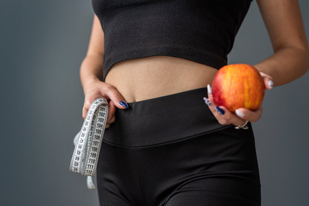 Trainer holding green apple and measuring tape for measuring body in fashionable sportswear on gray background isolated. healthy lifestyle conceptの写真素材