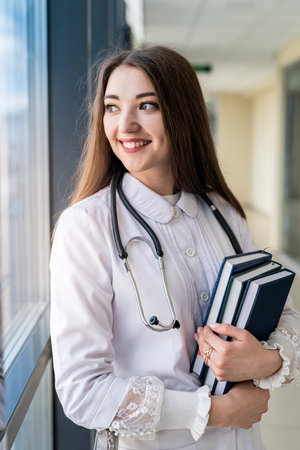 Vertical portrait of smiling young female doctor in uniform with stethoscope holding medical book while standing and looking away. concept of medicineの写真素材
