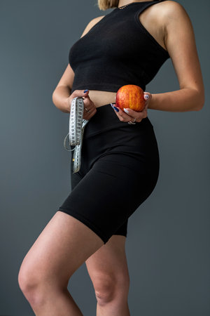 Young strong sports fitness trainer instructor woman wearing sports suit holding apple measuring tape isolated on gray background. Sport workout concept. healthy body conditionの写真素材
