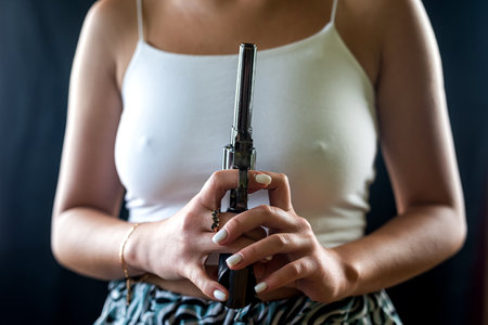 serious woman in stylish clothes posing faceless with revolver gun isolated on black background....