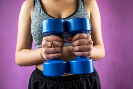 Portrait of cropped young attractive fitness woman holding dumbbells in studio on monochrome...