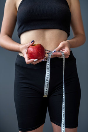 Trainer holding green apple and measuring tape for measuring body in fashionable sportswear on gray background isolated. healthy lifestyle conceptの写真素材