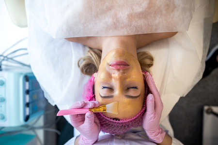 A beautician applies a gold mask to a client's face using an application brush. A beautiful woman lies and rests in a beauty salon. Anti-wrinkle spa treatments hydrate the face and even out the skin tone.の写真素材