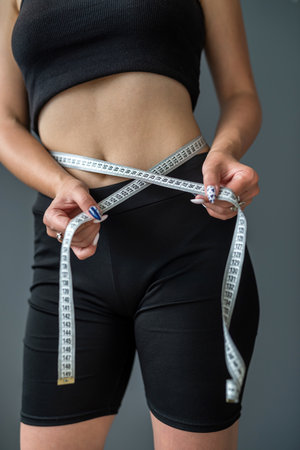 A young smiling woman measures her ideal waist with a tape measure and shows the result of her diet. Weight loss - portrait of a happy fitness girl. isolated body on gray backgroundの写真素材